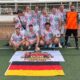 Mannschaftsfoto der DJK Bruchsal Old Boyz bei der Mallorca Football Tour. Die Spieler posieren in bunten Hawaiihemden vor einem Fußballtor. Im Vordergrund liegen ein Fußball sowie eine Banner mit dem Gruppenlogo und die deutsche Flagge.