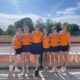 Sechs Frauen in orangefarbenen DJK-Tennisoutfits posieren lächelnd nebeneinander vor einem Sandplatz bei sonnigem Wetter.