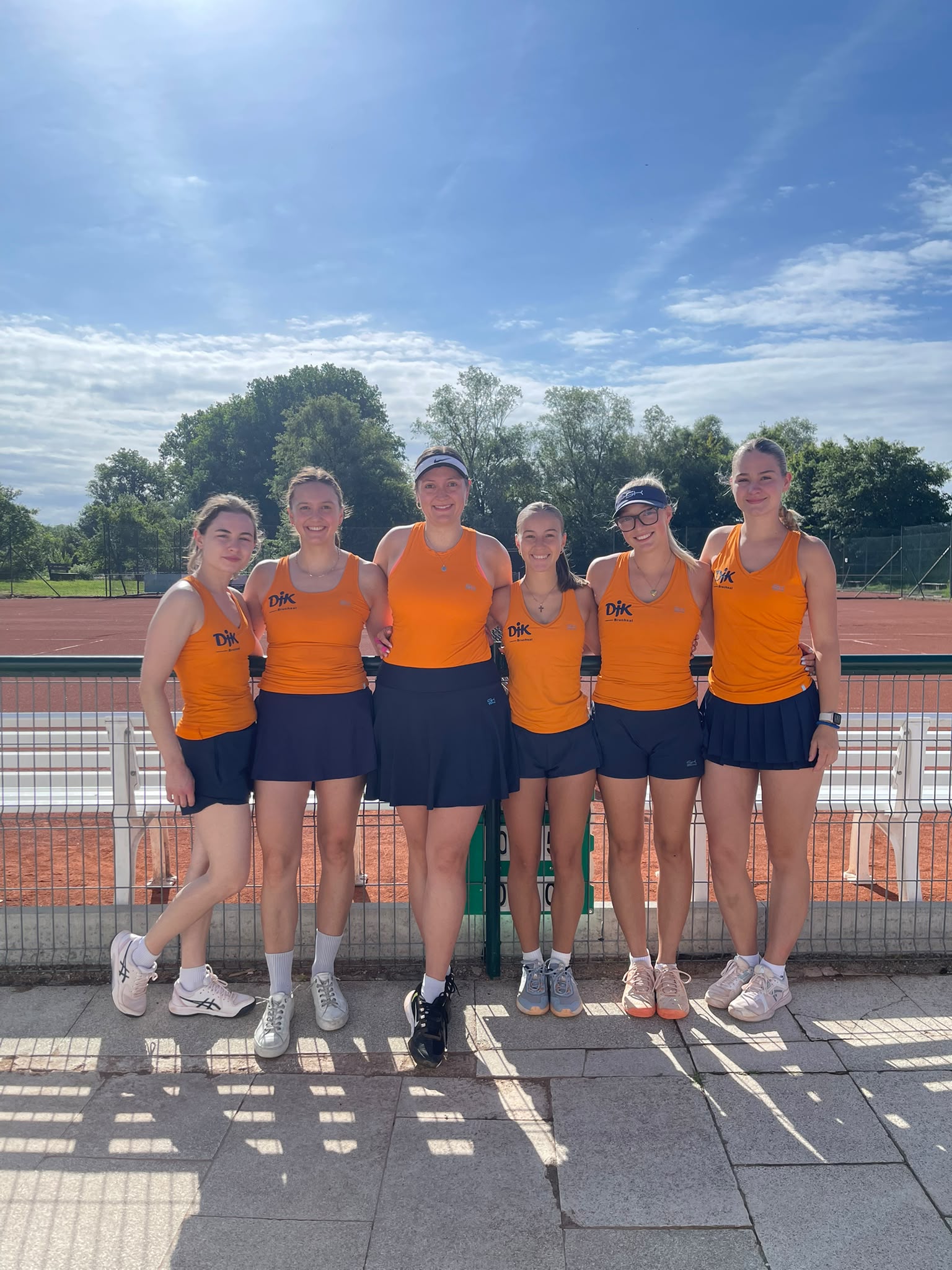 Sechs Frauen in orangefarbenen DJK-Tennisoutfits posieren lächelnd nebeneinander vor einem Sandplatz bei sonnigem Wetter.